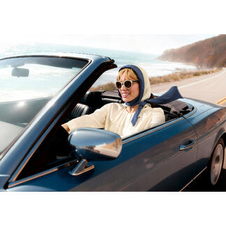 Femme dans une décapotable avec un foulard et des lunettes de soleil conduisant le long de la côte.