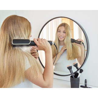 Femme avec une brosse à cheveux ionique devant un miroir dans une pièce moderne.