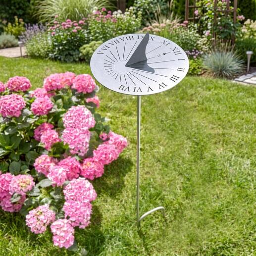 Cadran solaire en acier inoxydable sur une pelouse verte, entouré de fleurs roses.