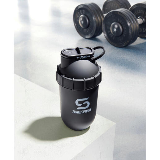 Ce shaker fitness utilise uniquement la force centrifuge créée par la forme arrondie de la capsule – sans aucun recoin dans lequel des résidus de poudre pourraient se coincer. Shaker noir sur une surface blanche avec des haltères en arrière-plan.