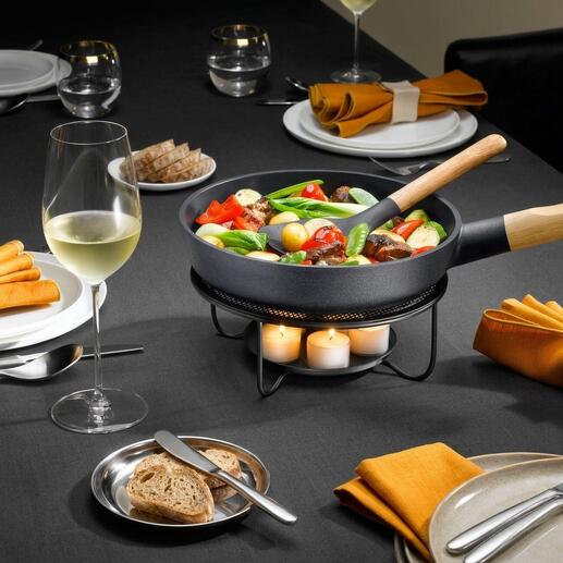 Poêle avec légumes sur un réchaud, à côté d'un verre de vin sur une table dressée.