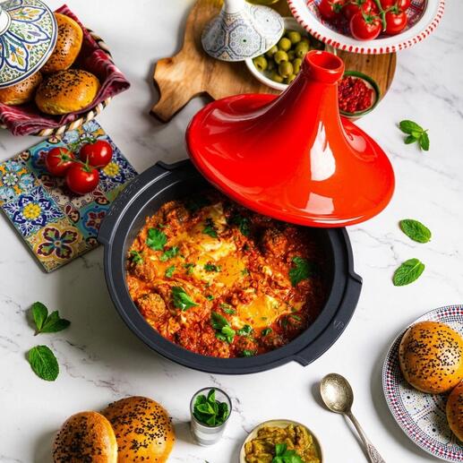 Tajine Peugeot Saveurs avec couvercle rouge, rempli de légumes colorés et d'herbes, sur une table avec des accompagnements.