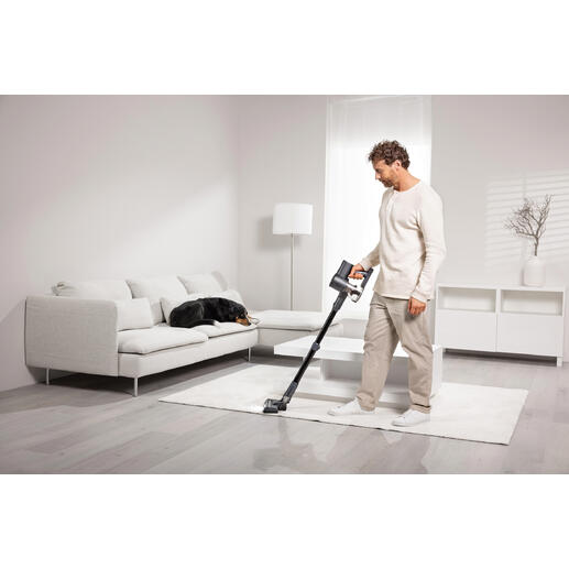 Un homme passe l'aspirateur sans fil sur un tapis, un chien est allongé sur un canapé.