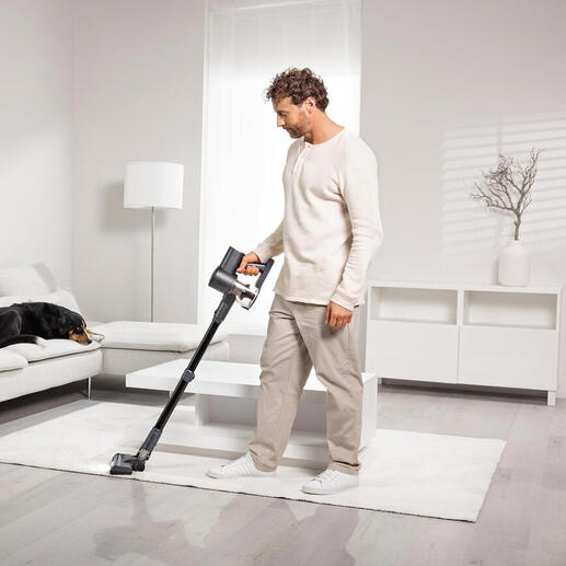 Un homme passe l'aspirateur sans fil sur un tapis, un chien est allongé sur un canapé.