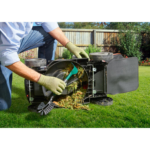 Personne nettoie une tondeuse à gazon retournée avec un outil, en enlevant les résidus d'herbe.