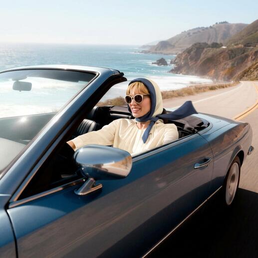 Femme dans une décapotable avec un foulard et des lunettes de soleil conduisant le long de la côte.