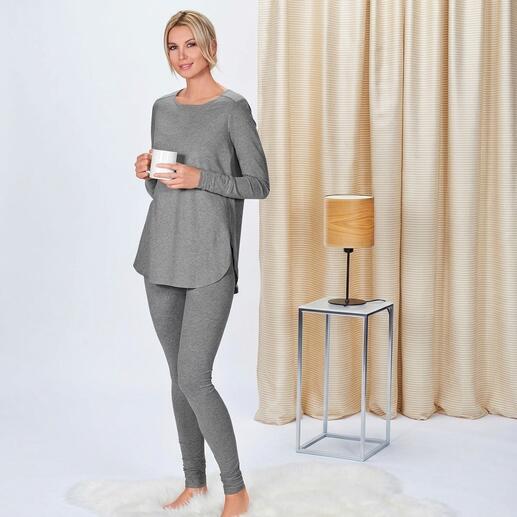 Femme portant un t-shirt long gris et un pantalon de jogging, tenant une tasse, debout dans un espace moderne.