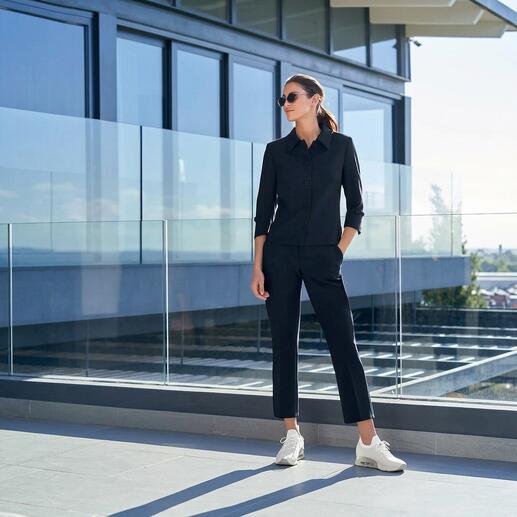 Femme en costume noir avec des baskets blanches se tient devant une balustrade en verre.