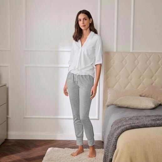 Femme en haut de pyjama blanc et pantalon de jogging gris debout à côté d'un lit.