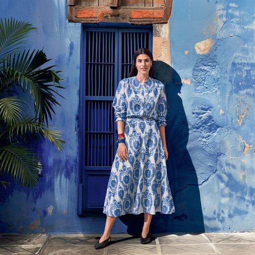 Femme portant une robe bleue à motifs blancs, se tenant devant un mur bleu.