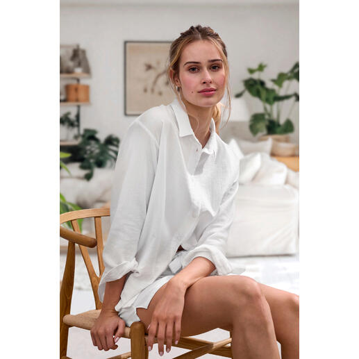 Blouse et Shorts Femme assise sur une chaise, portant une blouse en mousseline blanche Novila, avec des plantes naturelles en arrière-plan.