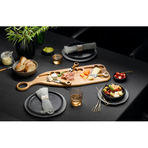 Planche de service design en bois d'acacia avec fromage, fruits et snacks sur une table sombre.