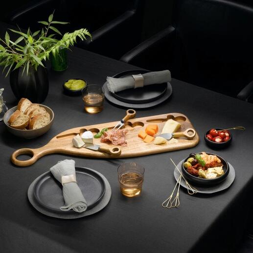 Planche de service design en bois d'acacia avec fromage, fruits et snacks sur une table sombre.