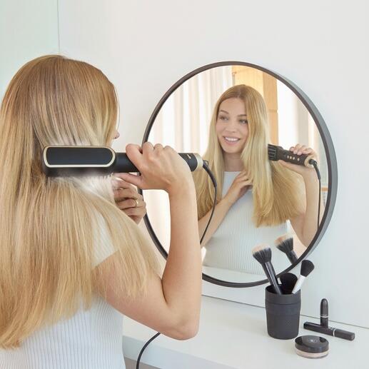 Femme avec une brosse à cheveux ionique devant un miroir dans une pièce moderne.