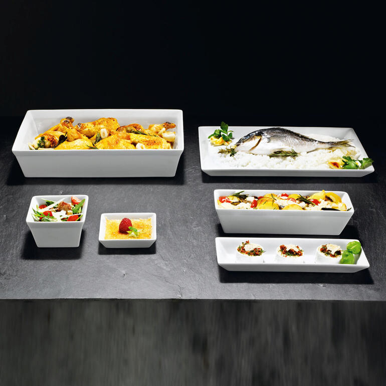 Différentes formes de vaisselle blanche sur une table sombre, avec différents aliments.