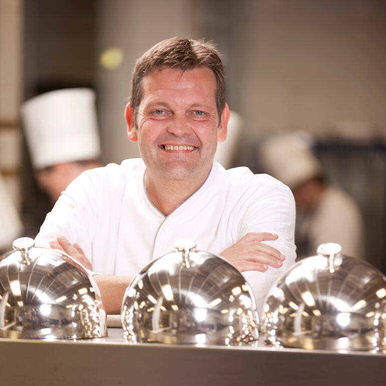 « Ma recommandation personnelle, pour tous les amateurs de Pro Idée » Cuisinier souriant à côté d'une table avec des couvercles en argent.