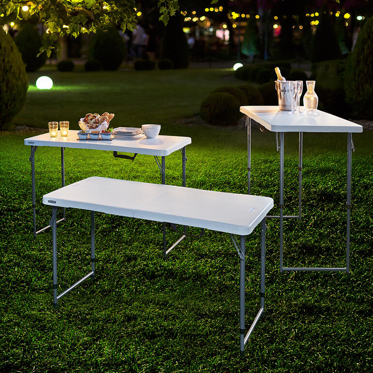 Table pliante réglable en hauteur Trois tables pliantes réglables en hauteur à l'extérieur, prêtes pour un pique-nique sous les lumières du soir.