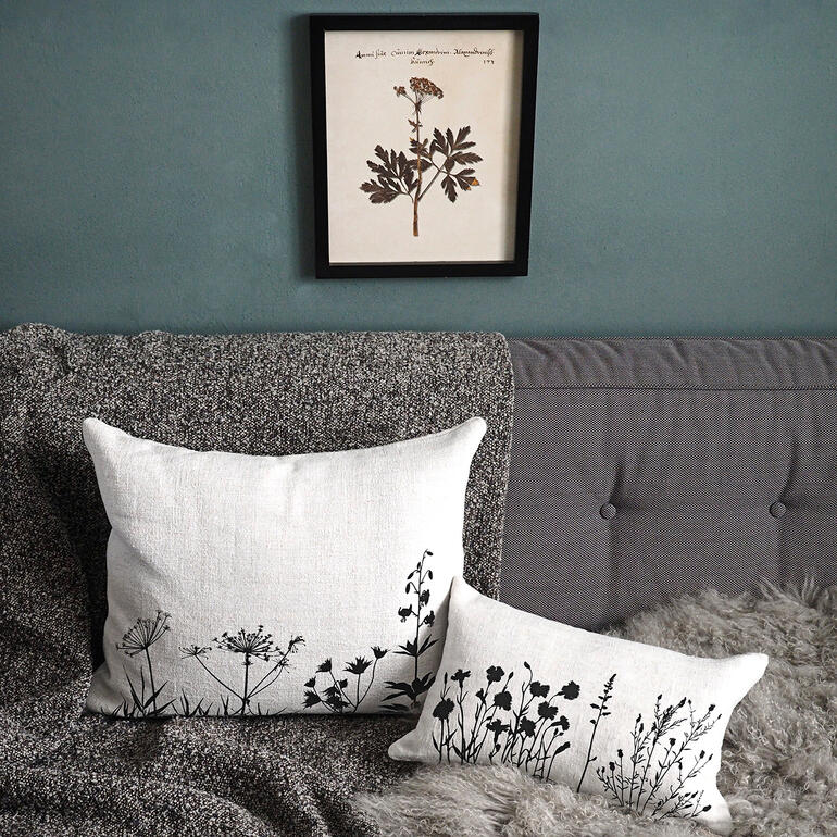Les imprimés aux motifs botaniques sont originaires d’une manufacture de Hanovre, qui réalise ses modèles de façon strictement artisanale. Deux coussins en lin avec des motifs de plantes noires sur un canapé, un tableau est accroché au-dessus.