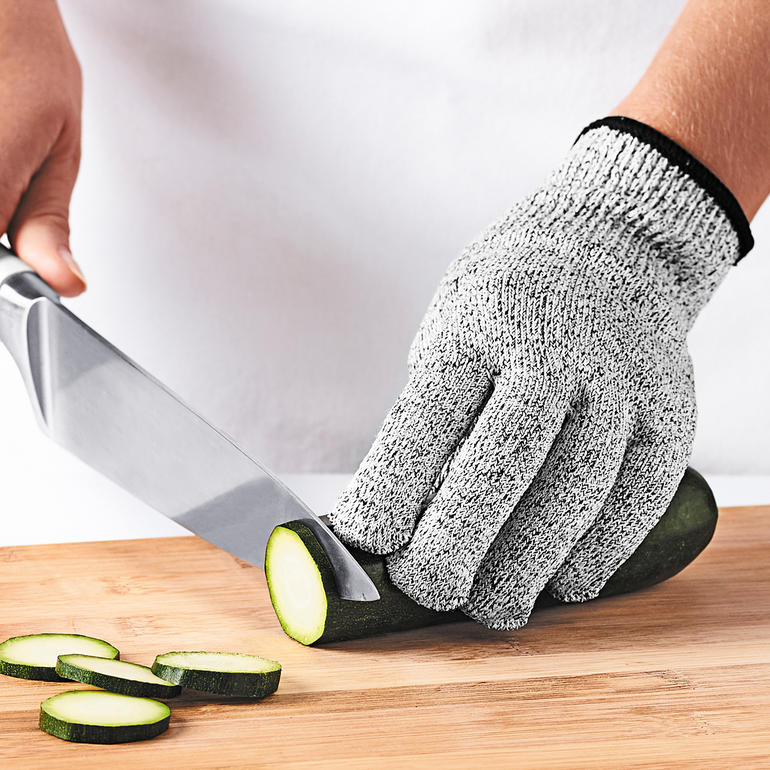 Main avec gant de protection Mastrad coupant un concombre sur une planche à découper.