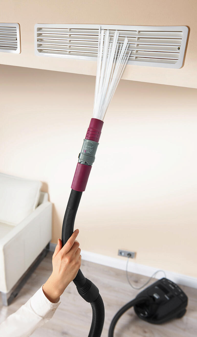 Main avec accessoire d'aspirateur nettoyant une grille de ventilation.