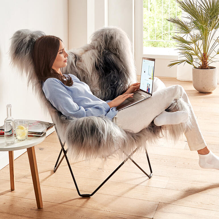 Le dossier du fauteuil affiche une largeur et une hauteur généreuses – pour un confort d’assise indiscutable, même pour les personnes de grande taille. Femme assise dans un fauteuil Mariposa avec un ordinateur portable, pièce lumineuse avec des plantes.