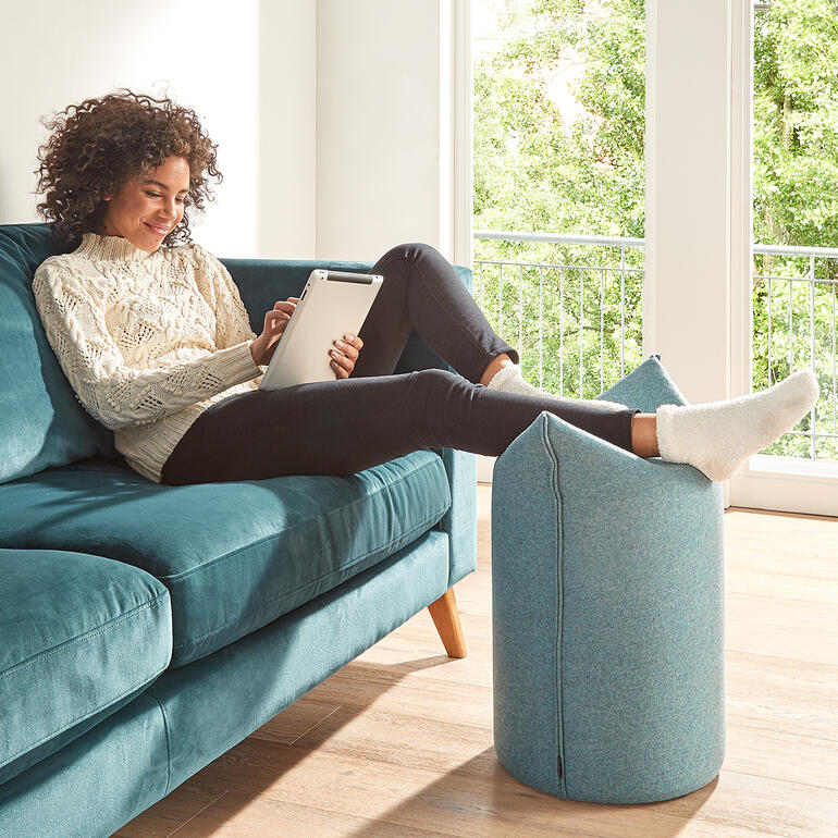 Également idéal pour y placer les jambes lors de vos instants de détente, pour regarder la télévision ou lire en toute quiétude. Femme assise sur un pouf bleu piolet utilisant une tablette.