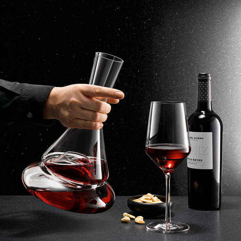 Décanter Doppio à deux niveaux avec du vin rouge à la main, à côté d'un verre à vin et des noix.