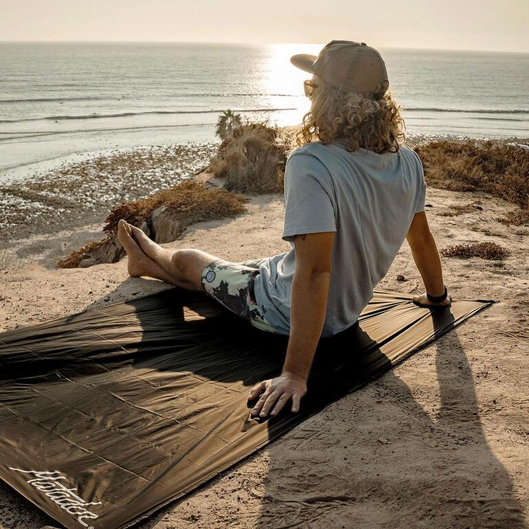 Homme assis sur une couverture ultralégère regardant la mer au coucher du soleil.
