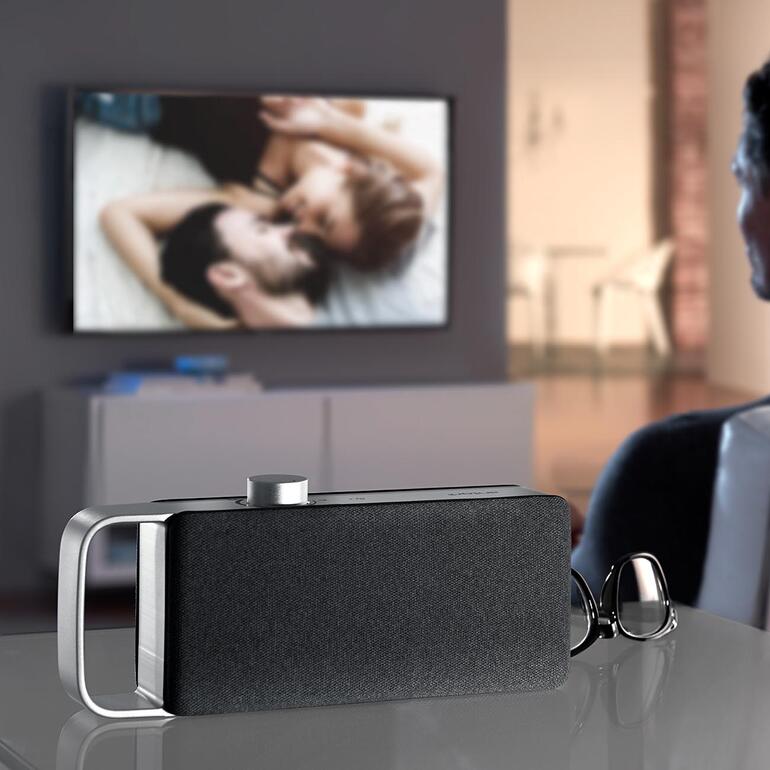 Amplificateur audio TV OSKAR, posé sur une table avec une télévision en arrière-plan.