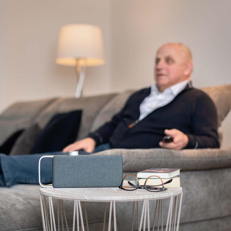 Un homme âgé assis sur un canapé, regardant la télévision et utilisant l'amplificateur audio TV OSKAR.