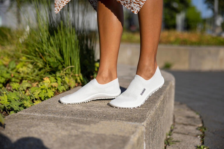 Chausson Barfuß-leguano® blanc sur le sol, avec une femme en arrière-plan.