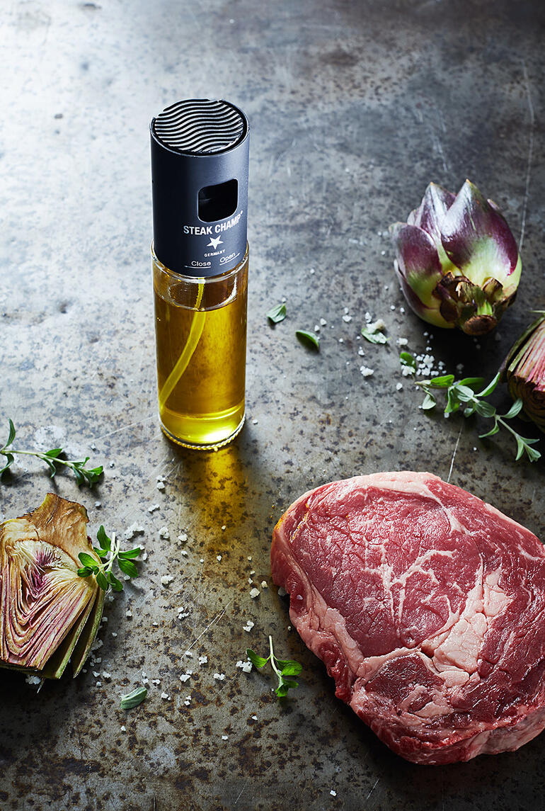 Vaporisateur d'huile avec filtre à côté d'un steak cru et des fleurs artificielles sur une table rustique.