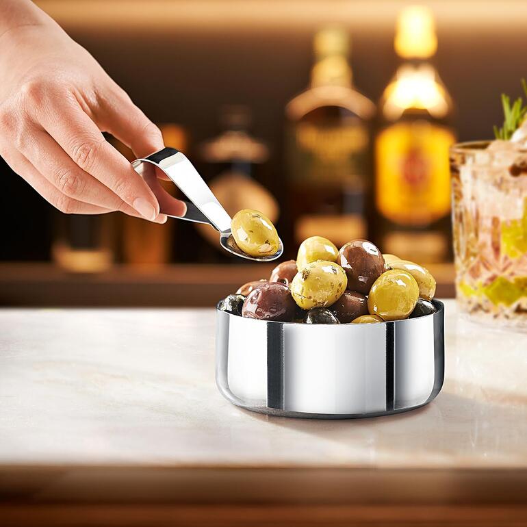 Bol à snacks design avec cuillère, rempli d'olives dans un environnement de bar.