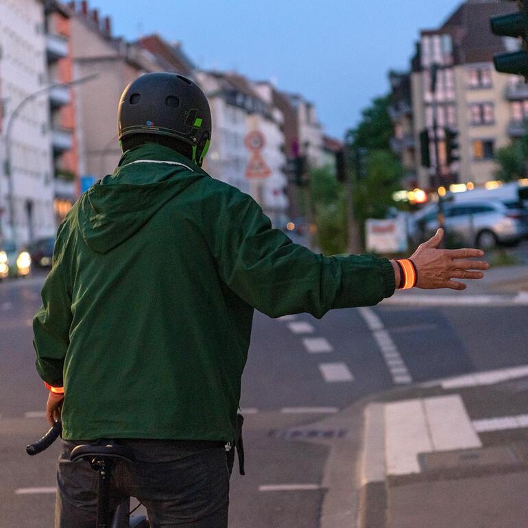 Grâce aux lumières LED supplémentaires sur le bracelet, vous serez mieux visible pour les autres usagers de la route, que ce soit au crépuscule ou dans lʼobscurité. Cycliste avec un bracelet lumineux faisant un signe de la main.