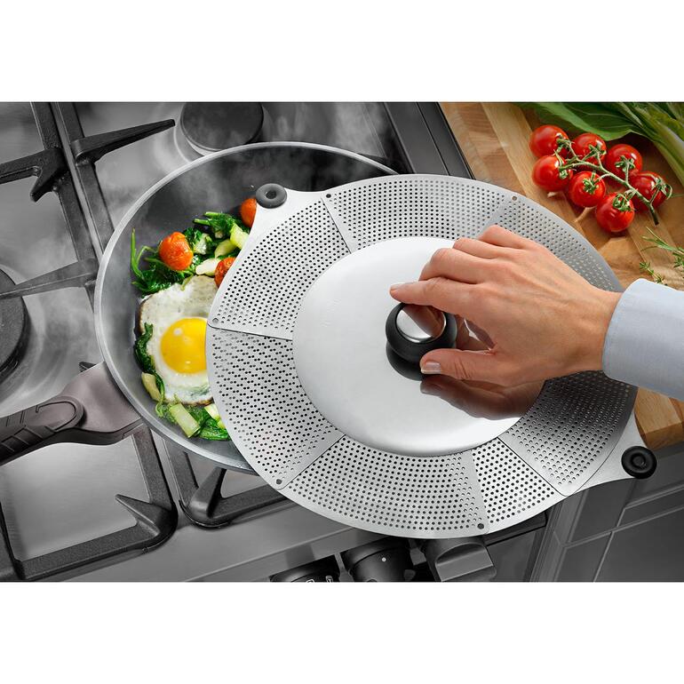 Main plaçant un couvercle anti-éclaboussures universel sur une casserole avec des légumes et un œuf sur une cuisinière