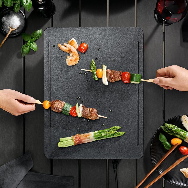 Grâce au système de chauffe innovant à 5 couches, la surface de cuisson est chauffée de manière optimale et uniforme. Mains mettant des brochettes de légumes et de viande sur le gril de table.
