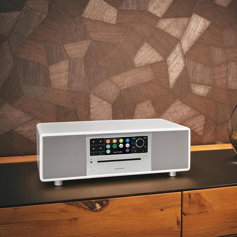 Sonoro PRESTIGE en blanc sur une table sombre devant un panneau en bois.