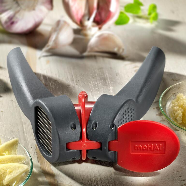 Éplucheur et presse-ail en noir avec un bouton rouge et des gousses d'ail en arrière-plan.