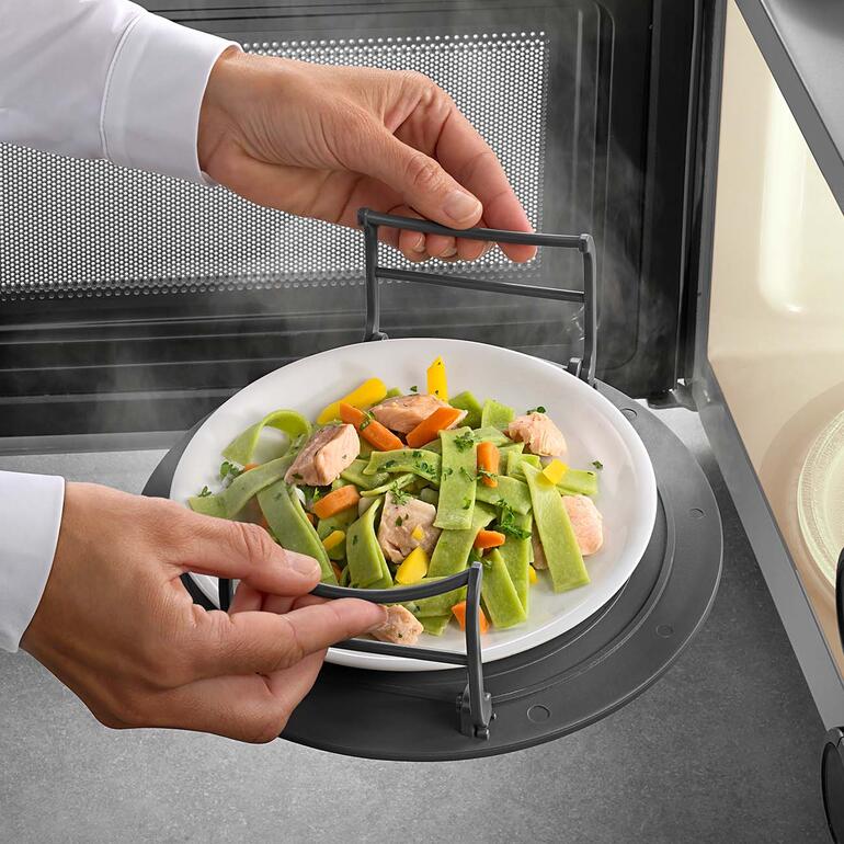 Idéale également en tant qu’aide au transport des assiettes chaudes. Personne tenant une assiette de légumes cuits à la vapeur tout en soulevant l'étagère à micro-ondes.