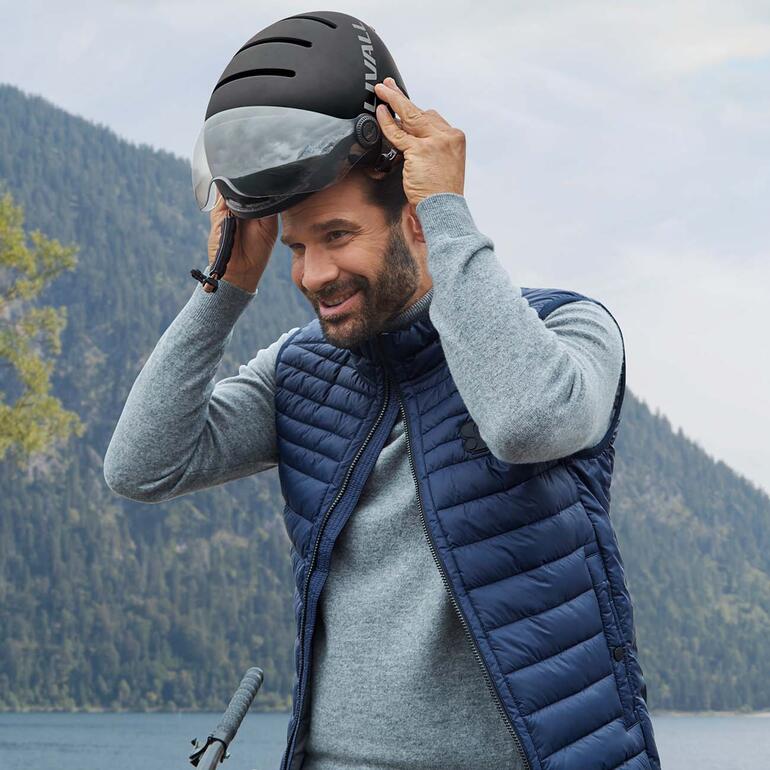 Le modèle à succès au design urbain stylé est désormais également équipé dʼune visière. Homme mettant son casque Livall L23 de vélo électrique au sommet de la montagne.