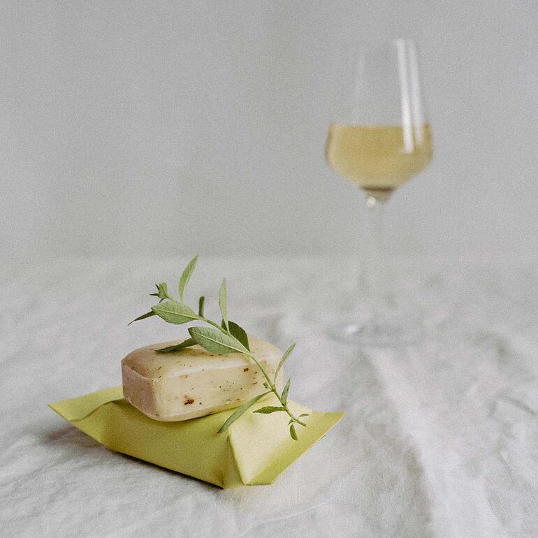 Savon The Real Wine Soap® décoré avec des feuilles et un verre de vin en arrière-plan.