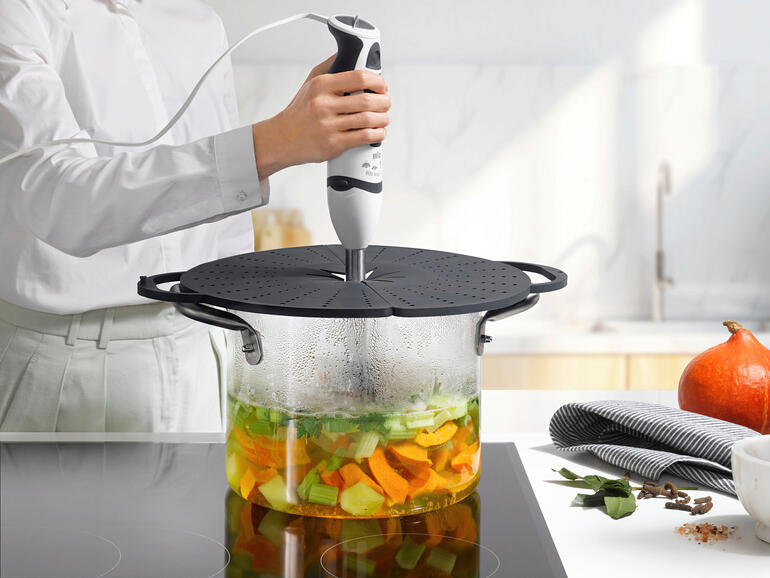 Avec ouverture flexible au centre pour un batteur ou un mixeur plongeant. Femme mixant des légumes dans une casserole avec un couvercle anti-éclaboussures en silicone.