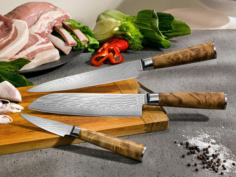 Trois couteaux en damas sur une planche à découper avec des ingrédients en arrière-plan