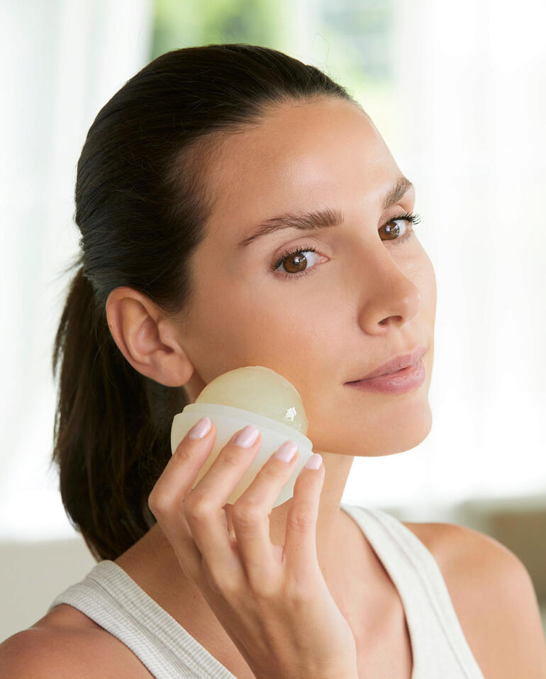 Enlevez la partie supérieure de la balle Cryo et passez lʼélixir glacé en le massant doucement par des mouvements circulaires sur le visage, le cou et le corps. Femme utilisant le Cryo-Elixir sur son visage.