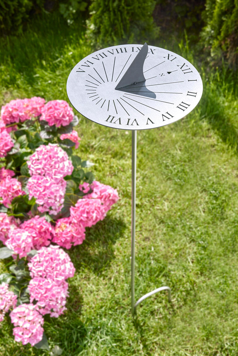 Cadran solaire en acier inoxydable sur une pelouse verte, entouré de fleurs roses.