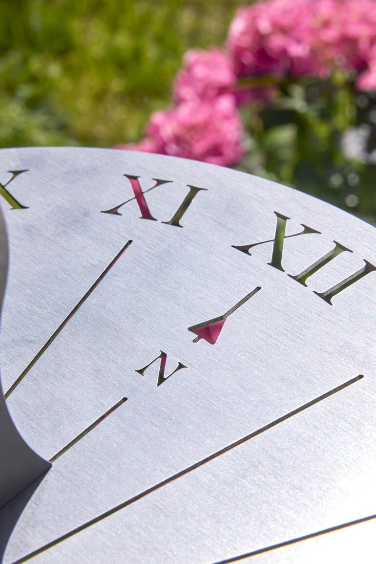 Gros plan des chiffres sur un cadran solaire en acier inoxydable avec des fleurs roses en arrière-plan.