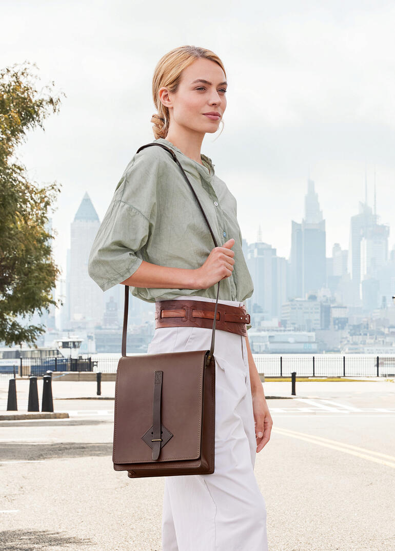 Femme portant un sac à carte Chiarugi marron sur l'épaule devant une vue de la ville.