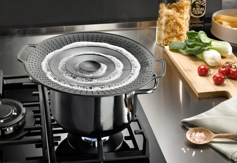 Casserole avec couvercle anti-éclaboussures sur la cuisinière, pâtes et légumes à côté.