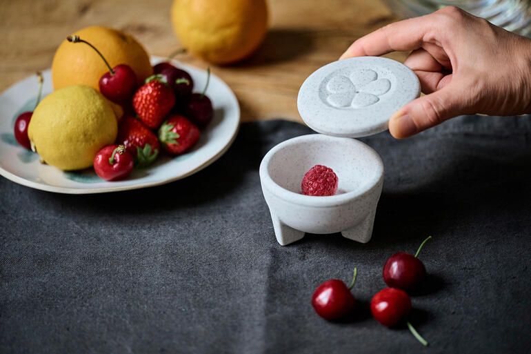 Piège à mouches en porcelaine avec une main ouvrant le couvercle et des fruits sur la table.