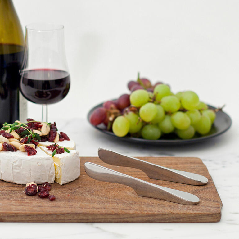 Ensemble de deux couteaux à tartiner flottants sur une planche en bois avec du fromage et des raisins.